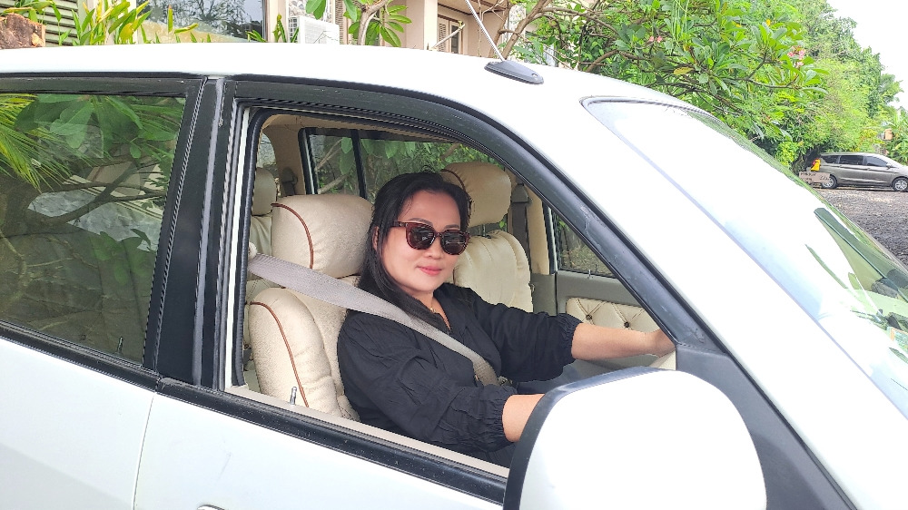 Professional Balinese lady driver in traditional dress standing beside a clean vehicle, with tropical palm trees and temple architecture in the background.